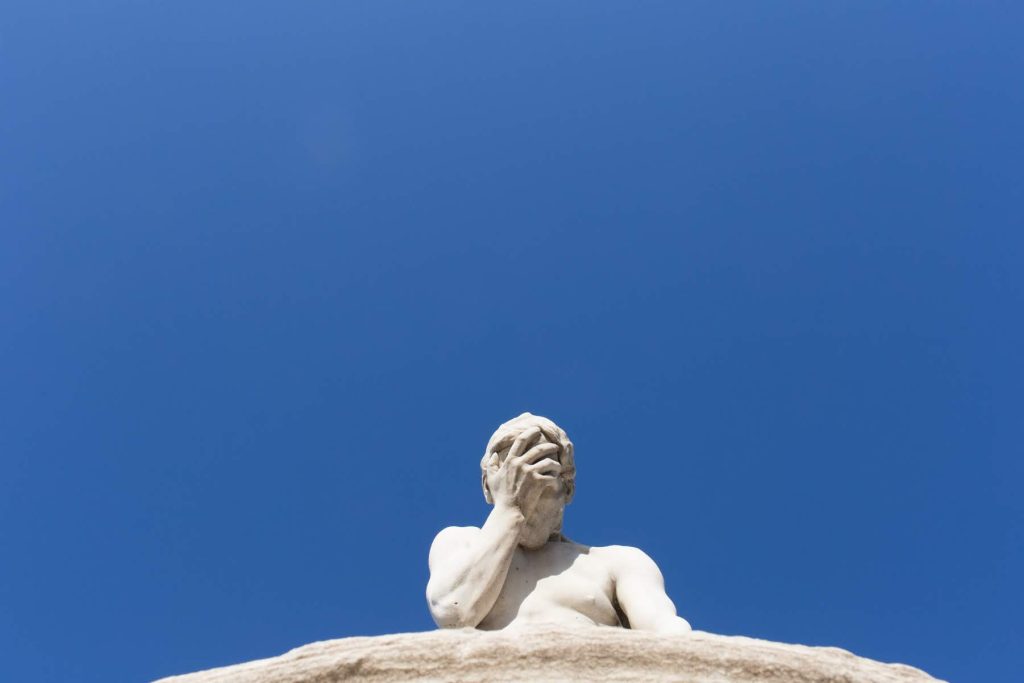 A marble statue photographed from below against a clear blue sky, one hand covering its face.