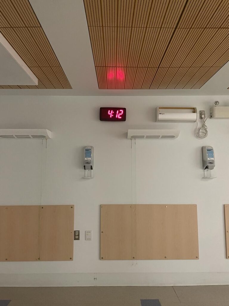 A hospital wall at night, a red digital clock reading 4:12 above two hand sanitizer dispensers and pale wood panels.