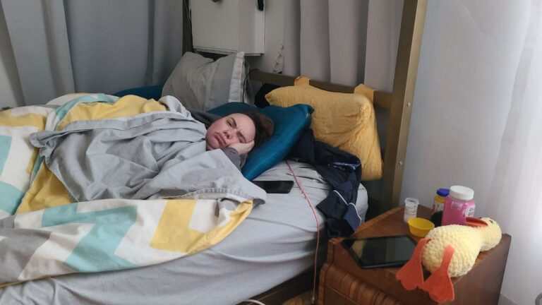A person lying in bed under a grey sheet and a striped blanket, head propped on one hand, a tablet, two bottles and a crocheted duck on the bedside table.