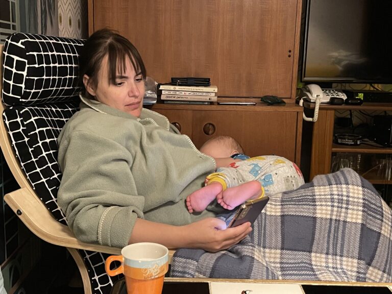 A parent reclining in an armchair nursing a baby, a phone in one hand and a mug on the table beside them.